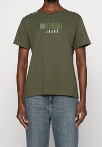 T-shirt en coton vert olive avec des manches courtes, présentant le texte "MOSH JEANS" en lettres grasses dans des teintes plus claires. Design simple et coupe décontractée.