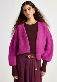 Blød pink cardigan med vide ærmer og åben front, kombineret med en brun top. Brun bælte med spænde fremhæver det lilla skørt.