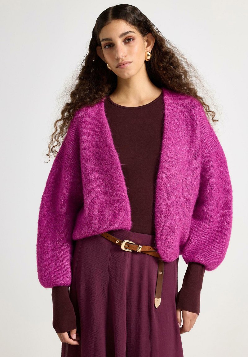 Blød pink cardigan med vide ærmer og åben front, kombineret med en brun top. Brun bælte med spænde fremhæver det lilla skørt.