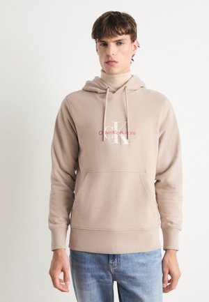 Sudadera beige de tejido suave con un bolsillo tipo canguro, que presenta un logo blanco "CK" y el texto "Calvin Klein Jeans" en rosa.