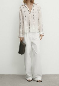 Camicia di lino beige chiaro con strisce verticali scure, abbinata a pantaloni bianchi a gamba larga e sandali neri; tiene in mano una borsa rettangolare marrone.