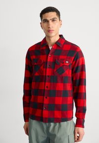Rød og sort ternet flannel skjorte med lange ærmer, to frontlommer og sorte knapper, båret over en hvid t-shirt.