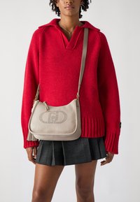 Bolso cruzado de cuero beige con detalle de logo perforado, herrajes dorados y un acabado texturizado, combinado con un suéter de punto rojo y falda de cuadros.