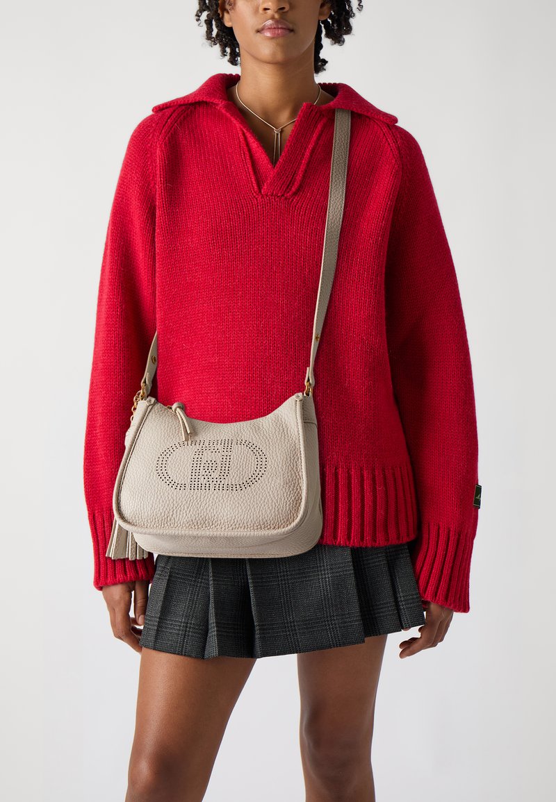 Bolso cruzado de cuero beige con detalle de logo perforado, herrajes dorados y un acabado texturizado, combinado con un suéter de punto rojo y falda de cuadros.