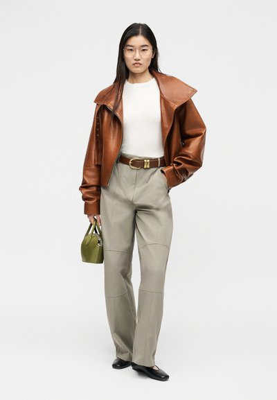 Femme portant une veste en cuir marron, une chemise blanche, un pantalon beige, des chaussures noires, des lunettes, tenant un petit sac à main vert, debout devant un fond blanc.
