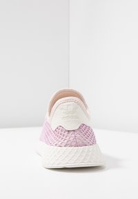 Jednoduchý růžový a bílý tenisky Adidas s prodyšným svrškem a texturovanou podrážkou, zobrazené ze zadní strany na bílém pozadí.