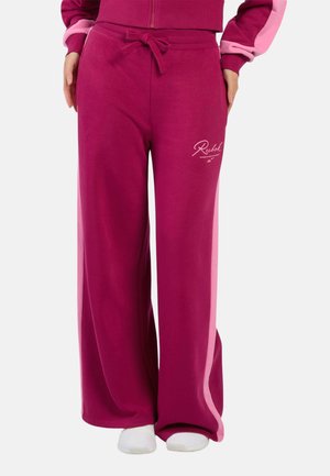 Vrouw draagt magenta wijde broek met roze zijstrepen, een taille met trekkoord en witte sneakers, staat met handen in de zakken.