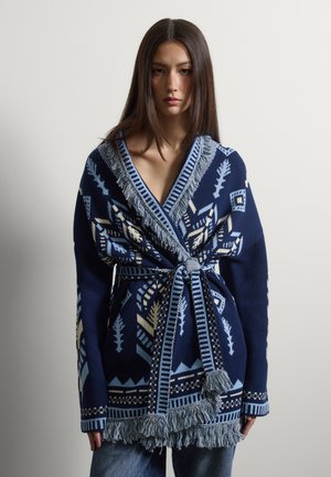 Donna che indossa un cardigan lavorato a maglia blu con motivo a fantasia, rifinito con frange e cintura annodata, in piedi davanti a uno sfondo grigio chiaro uniforme.