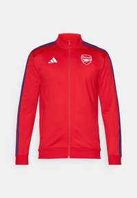 Rød lynlåsejakke med høj krave; har blå sidestriber og et hvidt Arsenal-logo på brystet. Fremstillet af glat, let materiale.