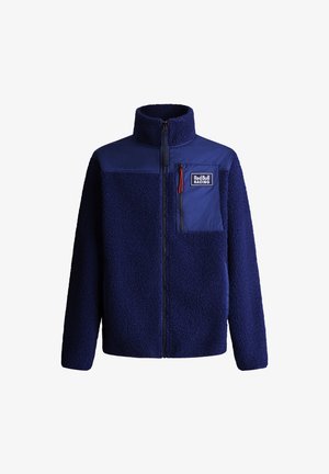 Tumesinine fliistek, millel on sile nailonist ülemine osa, kõrge krae, ettepaigutatav tõmblukk ja rinna tasku koos väikese punase tõmbluku tõmbega.