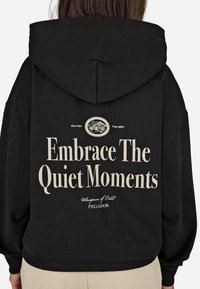 Μαύρη φούτερ με κουκούλα, με μεγάλη λευκή γραφική αναγραφή "Αγκαλιάστε τις Ήσυχες Στιγμές" στο πίσω μέρος και με έναν κυκλικό λογότυπο από πάνω.