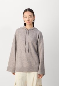 Sudadera con capucha de color beige claro, con mangas largas y dobladillo acanalado. Textura suave, diseño informal. Lleva puesta sobre pantalones cargo de color claro.