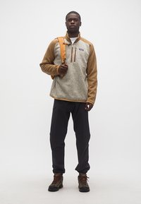 Άνδρας που στέκεται φορώντας ένα ταμπά και γκρι φλις πουλόβερ, μαύρο παντελόνι, παπούτσια πεζοπορίας και πορτοκαλί ιμάντα σακιδίου πλάτης πάνω σε έναν ώμο.