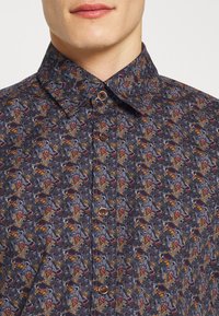 Homme portant une chemise à boutons sombre avec un motif floral dense dans des tons d'orange, de jaune, de violet et de gris.