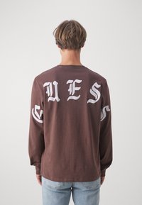 Maglietta a maniche lunghe marrone con grande testo gotico bianco sul retro e sulle maniche, con scollatura a girocollo e vestibilità comoda.