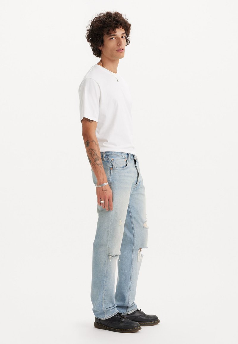 Maglietta bianca a maniche corte, jeans strappati azzurri chiari e scarpe nere stringate. Il modello si trova di lato, mostrando il taglio e le texture degli abiti.