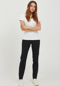 Young woman with long brown hair wearing a white short-sleeve top, black pants, white sneakers, standing with arms crossed on white background.