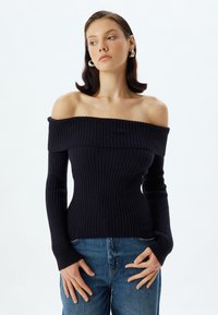 Marineblause, schulterfrei, mit langen Ärmeln, in figurnaher Silhouette und strukturiertem Stoff. Kombiniert mit blauen Jeans.