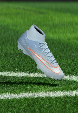 Chaussure de football Nike gris clair avec virgule orange flottant au-dessus de l’herbe verte avec des lignes blanches du terrain.