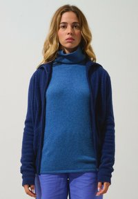 Blå turtlenecktröja under en marinblå zip-up jacka med ribbade manschetter. Outfitten är matchad med ljuslila byxor.