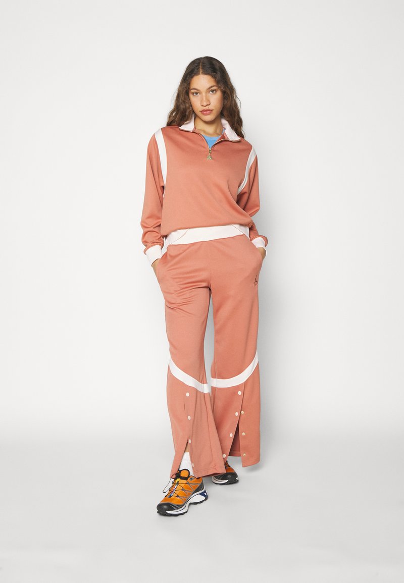 Ensemble de survêtement deux pièces en rose poussiéreux avec des accents blancs. Haut zippé, ample. Les pantalons ont des fentes latérales avec des pressions. Baskets orange et noires.