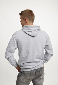 Uomo con capelli corti e castani che indossa un hoodie grigio chiaro e jeans neri sbiaditi, rivolto leggermente a sinistra su uno sfondo semplice.