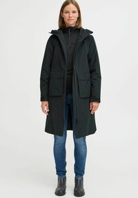 Black waterproof coat with a hood, zip closure, large front pockets, and a knee-length design, worn over a black top and blue jeans.