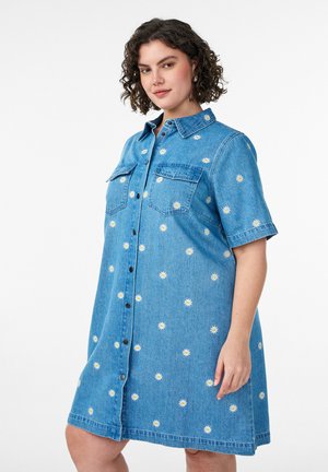 Kvinde med krøllet hår iført en blå denimkjole med hvide marguritprint, frontknapper og to brystlommer, stående foran en hvid baggrund.