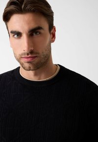 Zwarte gebreide trui met een geprofileerd geometrisch patroon, ronde hals en gedragen over een lichtbeige overhemd eronder.