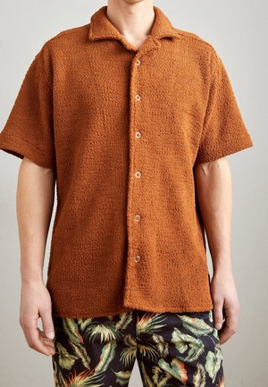 Mand iført en orange, struktureret kortærmet skjorte med knapper og mørke shorts med et stort grønt og orange tropisk bladmønster.