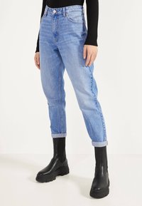 Ljusblå denimjeans med hög midja och upplagda vändor, tillsammans med höga svarta gummistövlar. Materialet verkar slätt med en klassisk design.