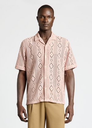 Homme à la peau foncée portant une chemise rose à manches courtes à motifs et un pantalon beige, debout devant un fond gris clair uni.
