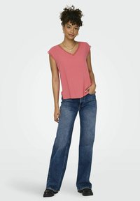 Rosa ermeløs topp med V-hals og blonder, parret med vide blå denimjeans og svarte sko, stående mot en lys bakgrunn.