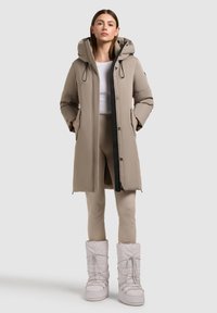 Beige padded parka with a hood, black zipper, and side pockets, paired with light-colored fitted leggings and beige snow boots. Smooth texture.