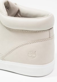 Talon de sneaker en cuir beige clair avec col matelassé et logo arbre Timberland embossé au-dessus de la semelle en caoutchouc blanche.
