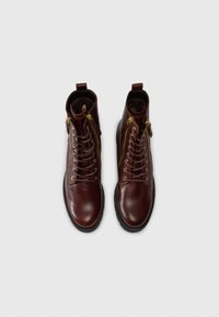 Botas de Tobillo de cuero marrón con una superficie lisa, con cordones en el frente y dos cremalleras laterales. Presenta punta redonda y suela de goma texturizada.