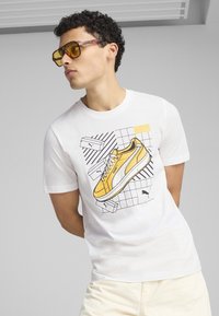 Camiseta blanca con un gráfico de una zapatilla amarilla, con contornos negros, sobre un fondo de cuadrícula. El diseño incluye ilustraciones adicionales de cajas de zapatos.