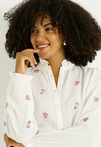 Camisa blanca de botones con bordado floral en rosa y azul. Corte holgado con cuello con volantes y tejido texturizado. Mangas largas fruncidas.