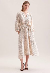 Hellbeiges Kleid mit grauen Paisley-Mustern, V-Ausschnitt, langen Ärmeln, Taillengürtel, Seitentaschen, fließendem Stoff, knöchellang, getragen mit schwarzen Sandalen.
