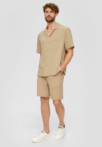 Lichtbeige korte mouwen shirt en bijpassende shortset, gemaakt van textuurstof, met een knoopsluiting aan de voorkant en een borstzak. Witte sneakers.