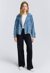 Svetlo modra jeans jakna z širokim ovratnikom in dolgimi rokavi, kombinirana s temnimi jeans hlačami in bež čevlji, na nevtralnem ozadju.