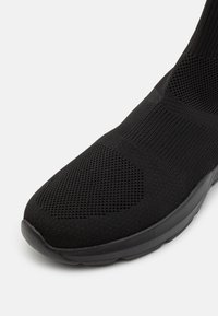 Černá sportovní obuv s pleteným svrškem, který má texturovaný vzor, polstrovaný límec a flexibilní gumovou podrážku.