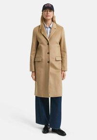 Beige wool coat with a tailored design, featuring a buttoned front, two side pockets, and a seamless finish. Paired with wide-legged denim pants.