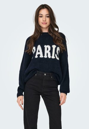 Granatowy sweter z dzianiny z białym napisem "PARIS", szerokimi rękawami i luźnym krojem, w połączeniu z czarnymi dżinsami. Gładka tekstura i casualowy design.
