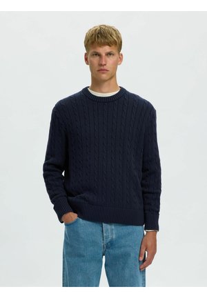 Jeune homme portant un pull en maille torsadée bleu marine et un jean bleu clair, debout devant un fond uni clair, les mains partiellement dans les poches.