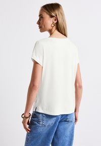 Bílý top s krátkým rukávem z hladké tkaniny, s kulatým výstřihem a bočními rozparky; kombinovaný se světle modrými džínami. Minimalistický design.