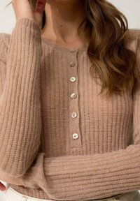 Beżowy kardigan z prążkowanego materiału, z okrągłym dekoltem, długimi rękawami i zapięciem na guziki w jasnoróżowym kolorze. Miękka faktura i dopasowany krój.