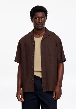 Homme aux cheveux courts et bouclés portant une chemise à manches courtes marron foncé boutonnée par-dessus un t-shirt beige et un pantalon foncé, debout sur fond blanc.