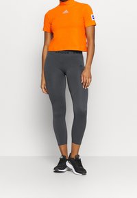 Pomarańczowy sportowy top z wysokim kołnierzem i logo Adidas; zestawiony z dopasowanymi szarymi legginsami z czarnym logo w pasie i teksturą.