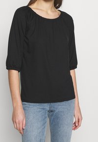 Kvinna i svart löst sittande blus med rynkad halsringning och ärmar, i kombination med ljusblå denimjeans, stående mot en enfärgad bakgrund.